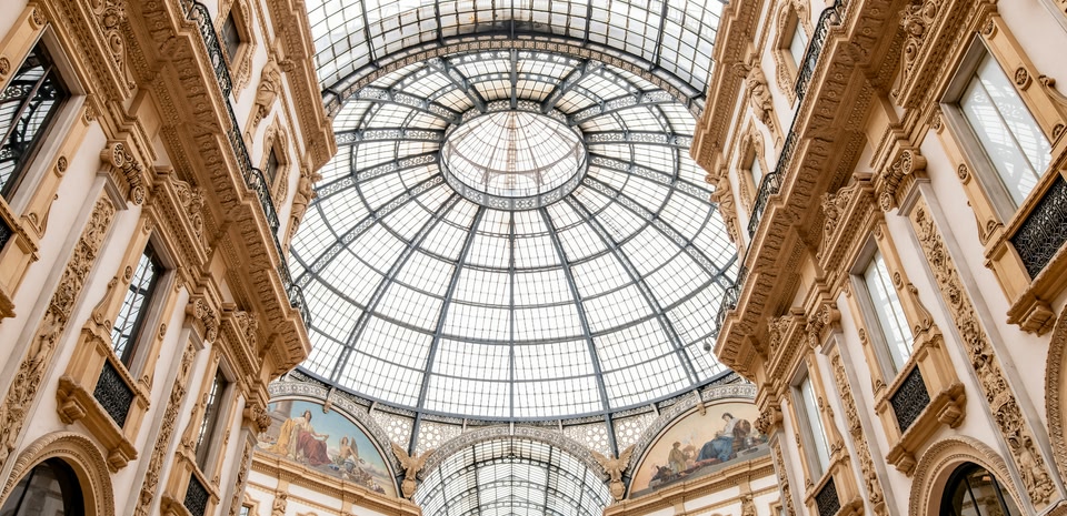 Techo de cúpula de vidrio ornamentado con estructura de hierro en una galería comercial histórica, con paredes decoradas en tonos crema y dorado y ventanas arqueadas.