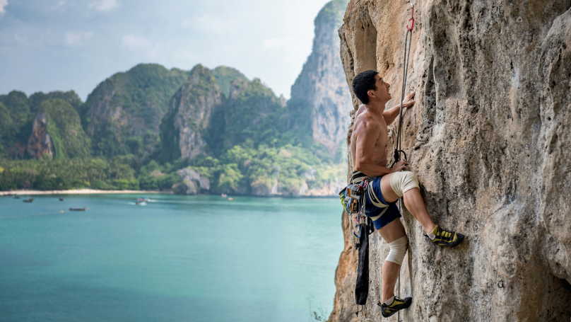 Eine atemberaubende Aufnahme eines mutigen Mannes, der eine felsige Klippe erklimmt, mit dem kristallblauen Meer und den majestätischen Bergen im Hintergrund.