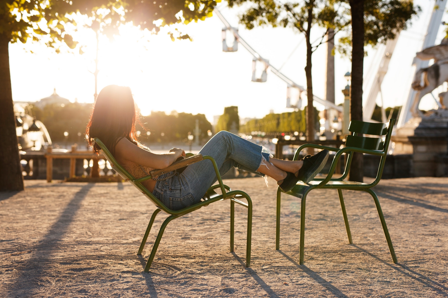 Young woman leaning against a chair, feet on another chair, watching a Ferris wheel at sunset.