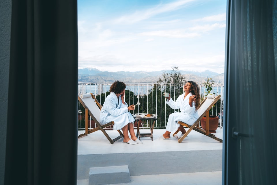 Two women sitting in dressing gowns on deckchairs on a balcony, overlooking a lake and mountains, enjoying coffee and talking