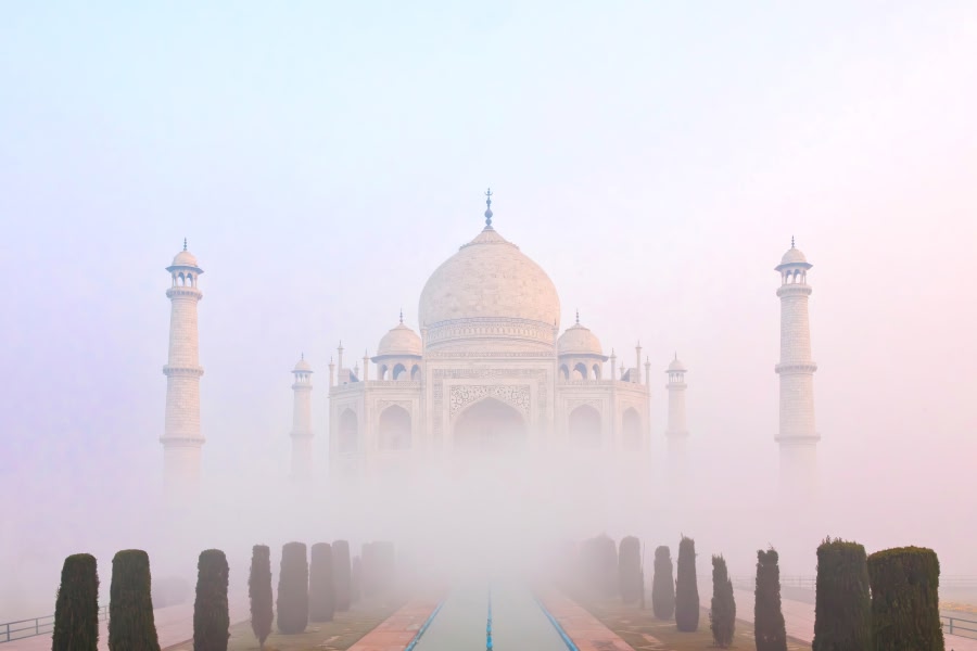 The Taj Mahal in Agra enveloped in soft morning mist, its white marble central dome and four soaring minarets emerging ethereally from the haze, with cypress trees lining the empty central garden pathway.