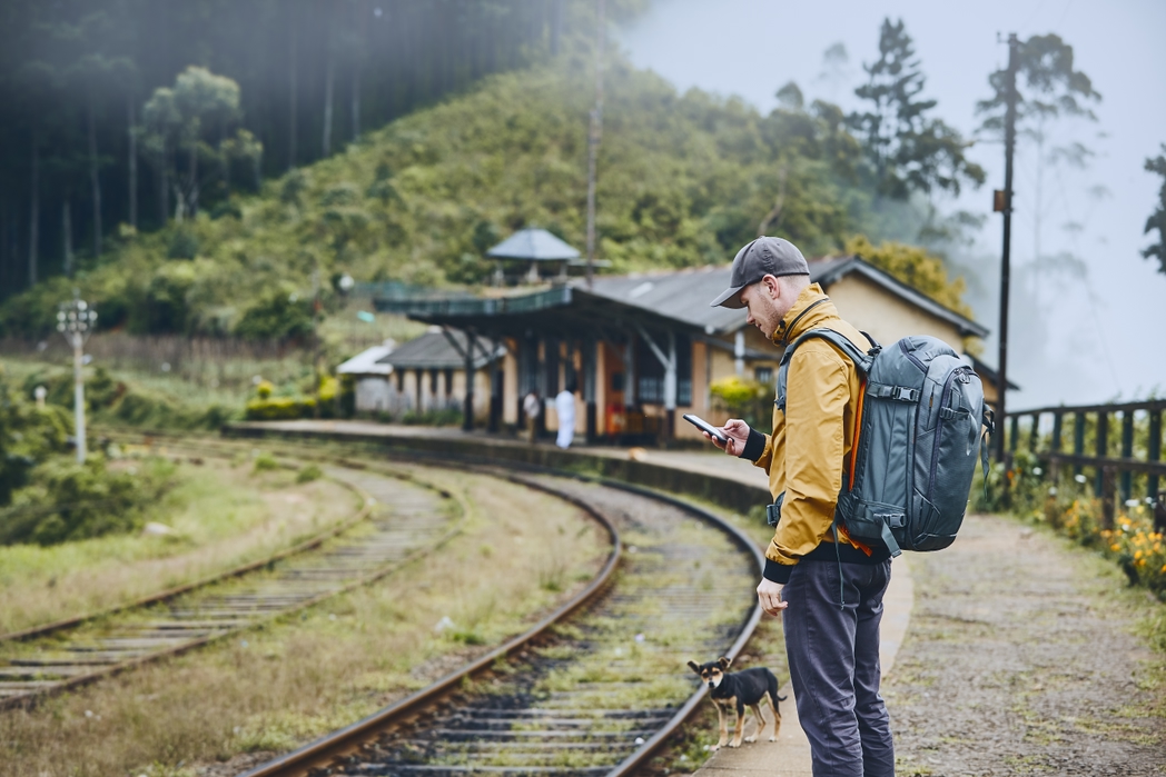Young tourist with backpack using phone and waiting for train. Mountains railway station in clouds, Sri Lanka.