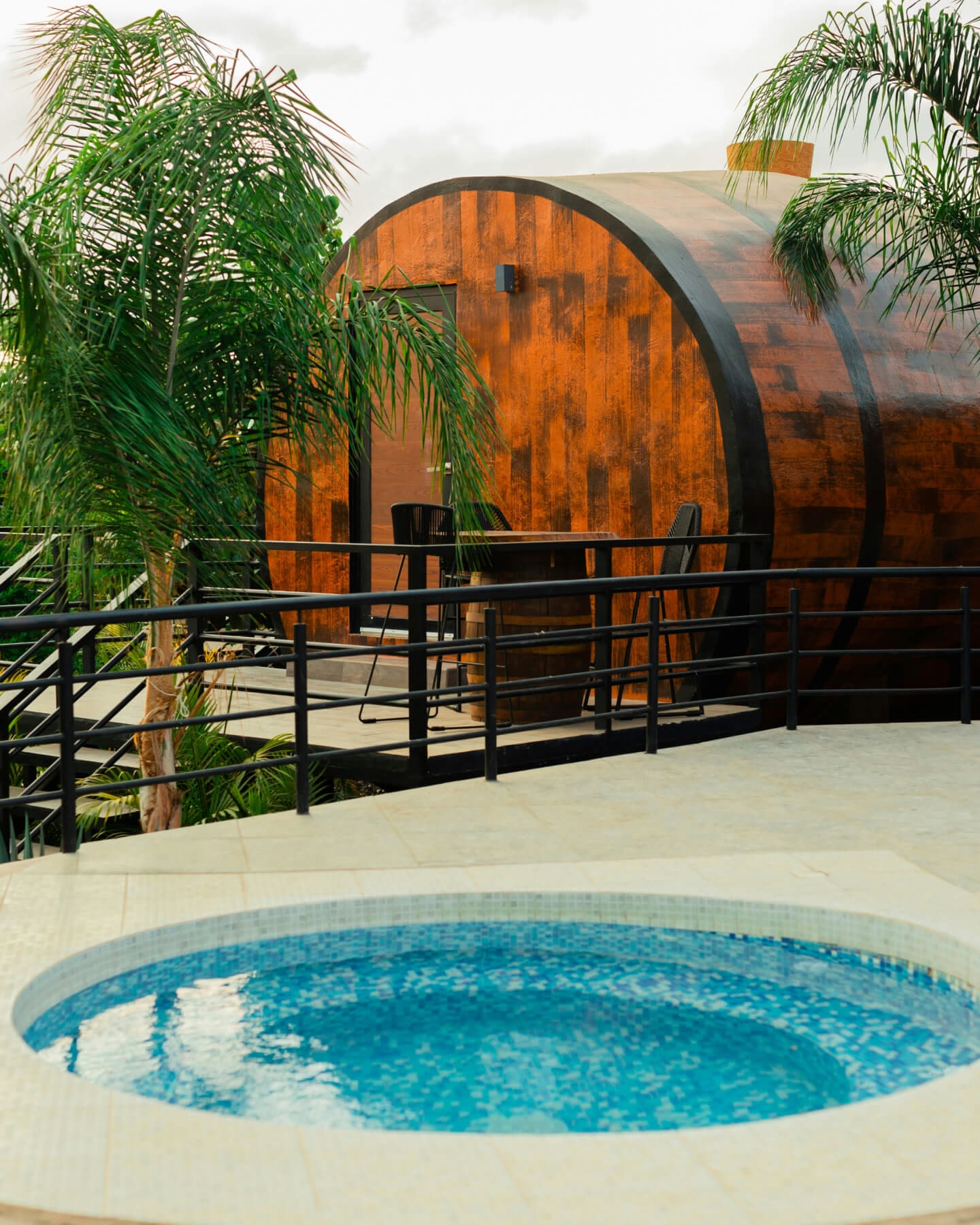 A barrel-style wooden cabin with a small hot tub in the foreground and palm trees on either side