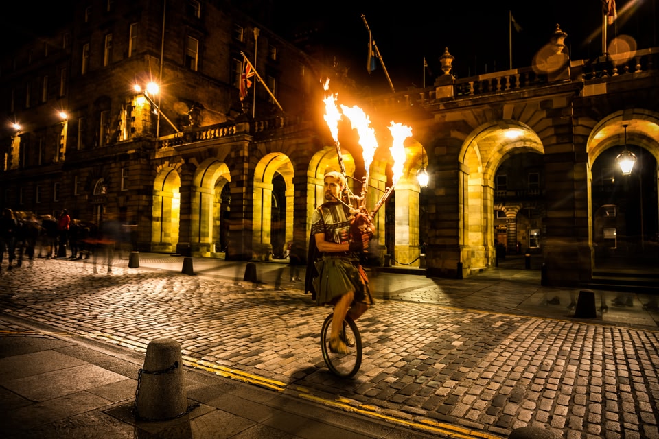 A bagpiper in costume riding on a unicycle beside the entrance to Lothian & Borders Police building on The Royal Mile in Edinburgh, Scotland, with flames coming out of the pipes.