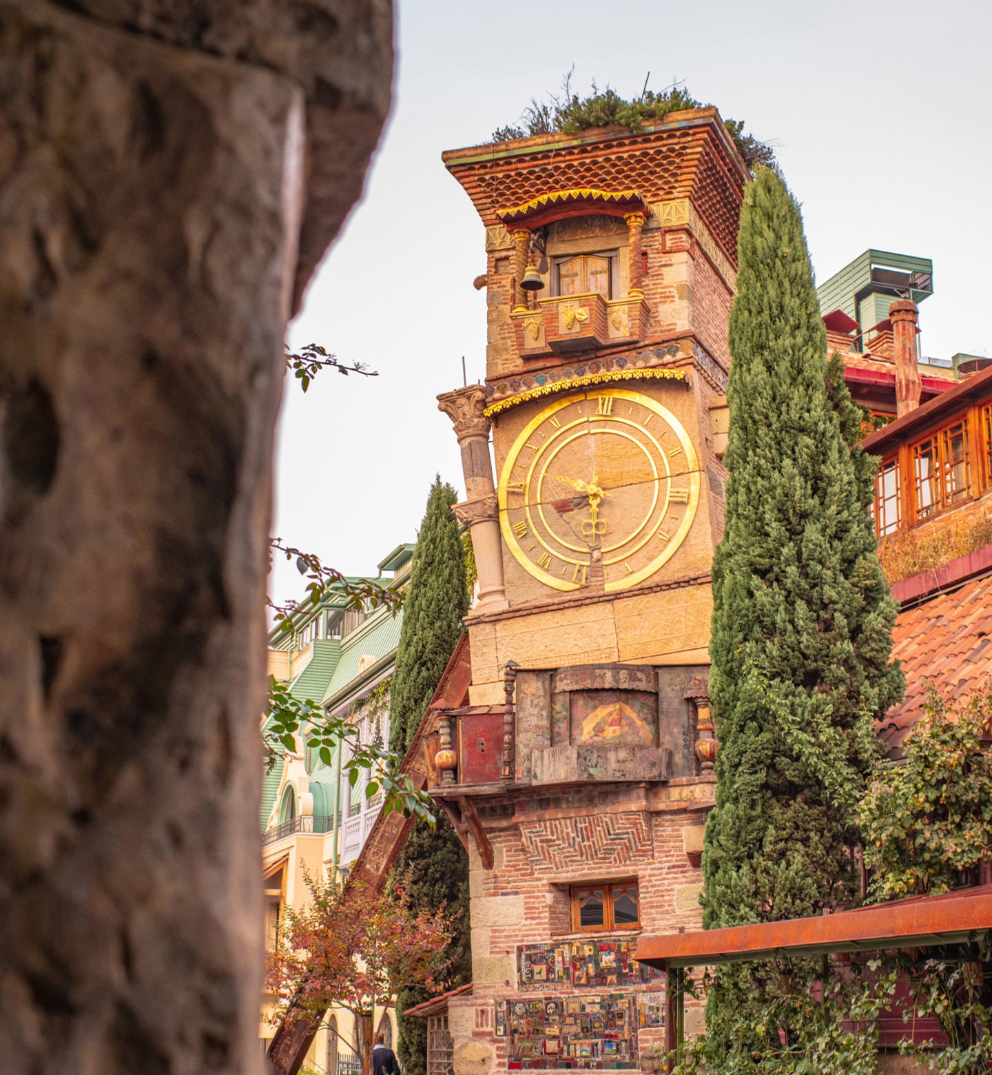 Close up shot of a quirky, leaning clock tower flanked by green trees in Tbilisi, Georgia.