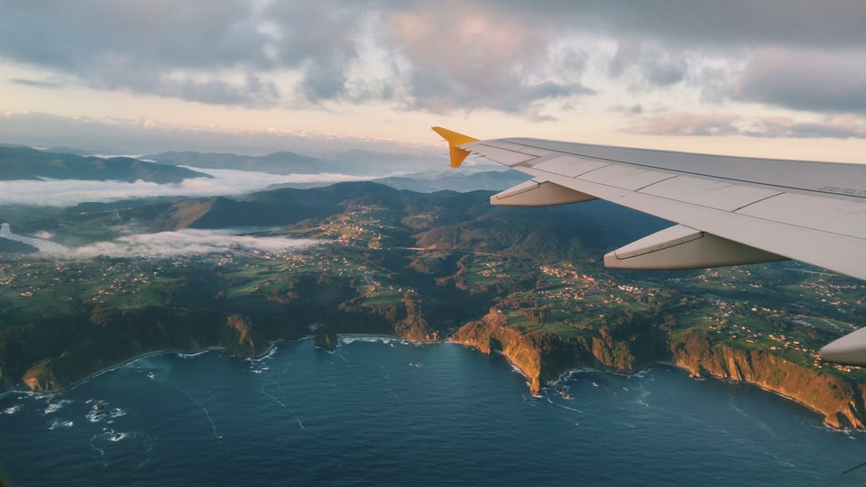 The left wing of a plane as it flies above a mountainous bay.