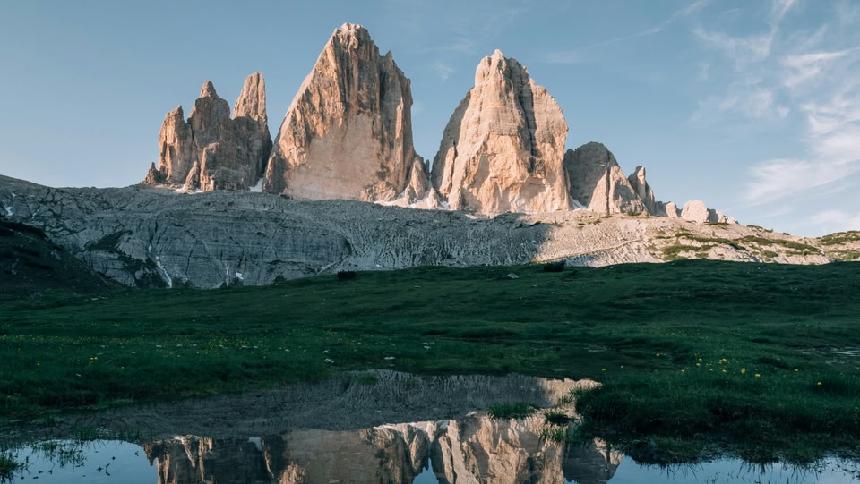 Paesaggio montano con le Tre Cime di Lavaredo sullo sfondo e un prato verde con un piccolo lago in primo piano.