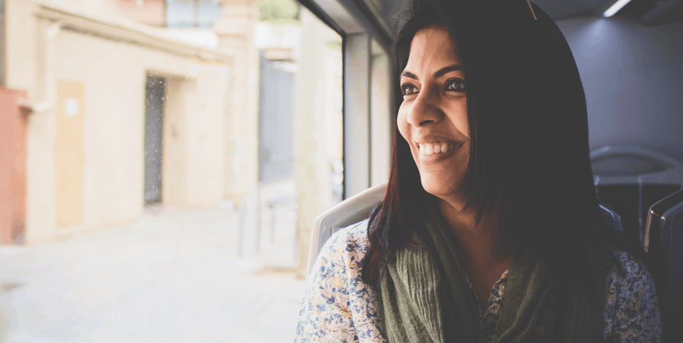 "A close-up of a cheerful woman with black hair sitting on a train, smiling warmly as she gazes out of the window.