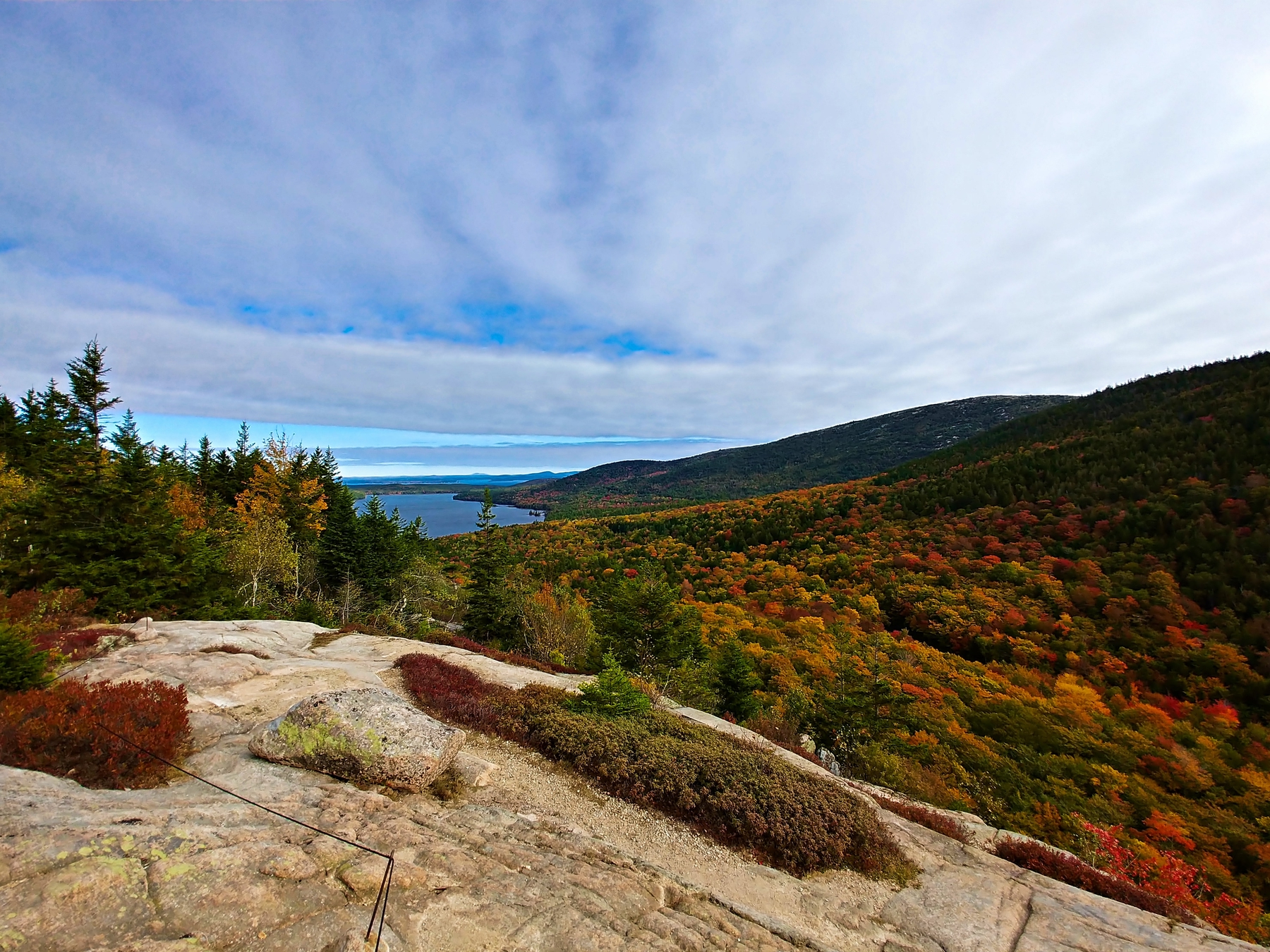 A brown cliff overlooks red, yellow, and orange trees with an ocean in the distance.