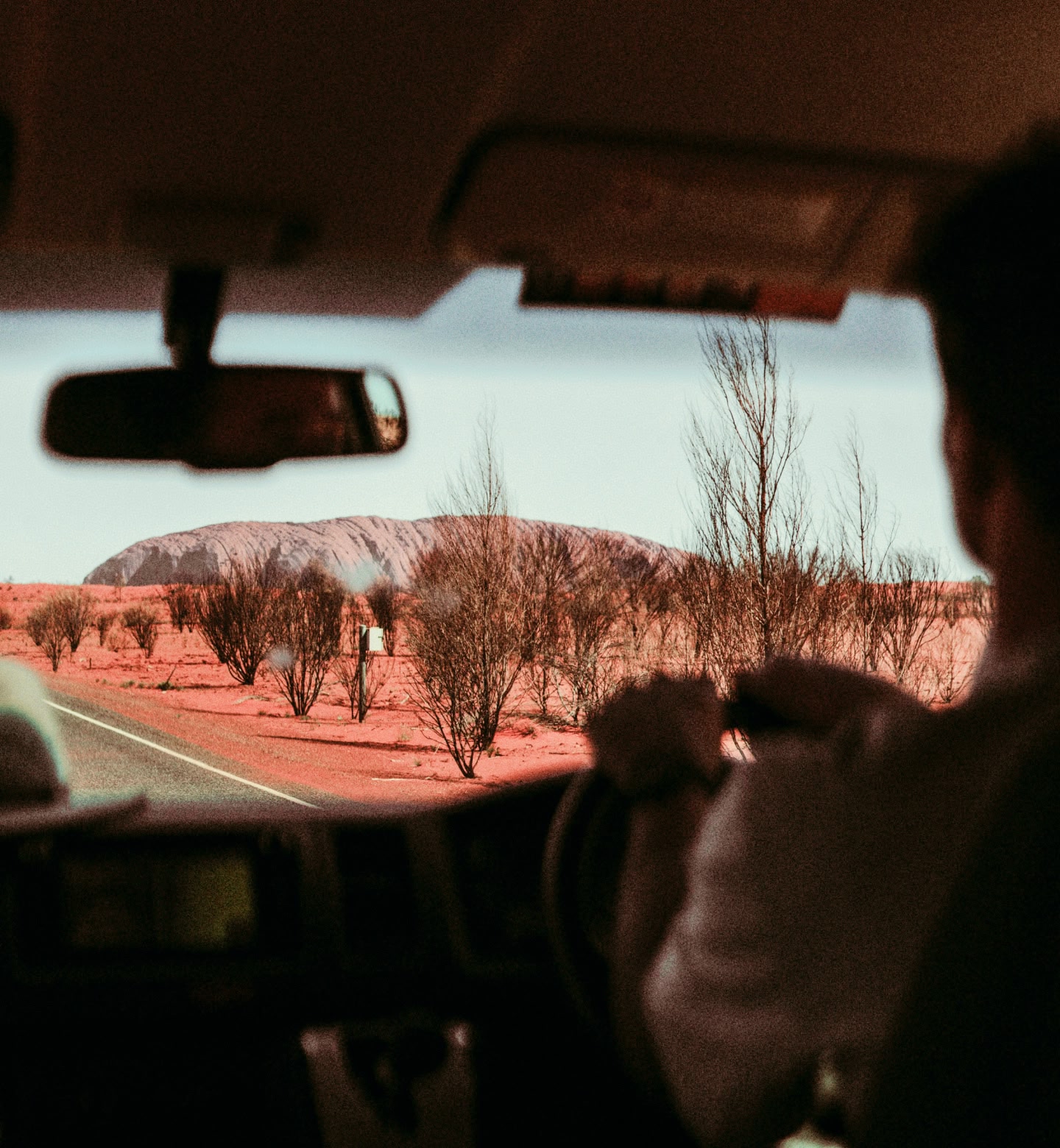 A man looks at a red landscape and Uluru rock, Australia, from behind the steering wheel of a car