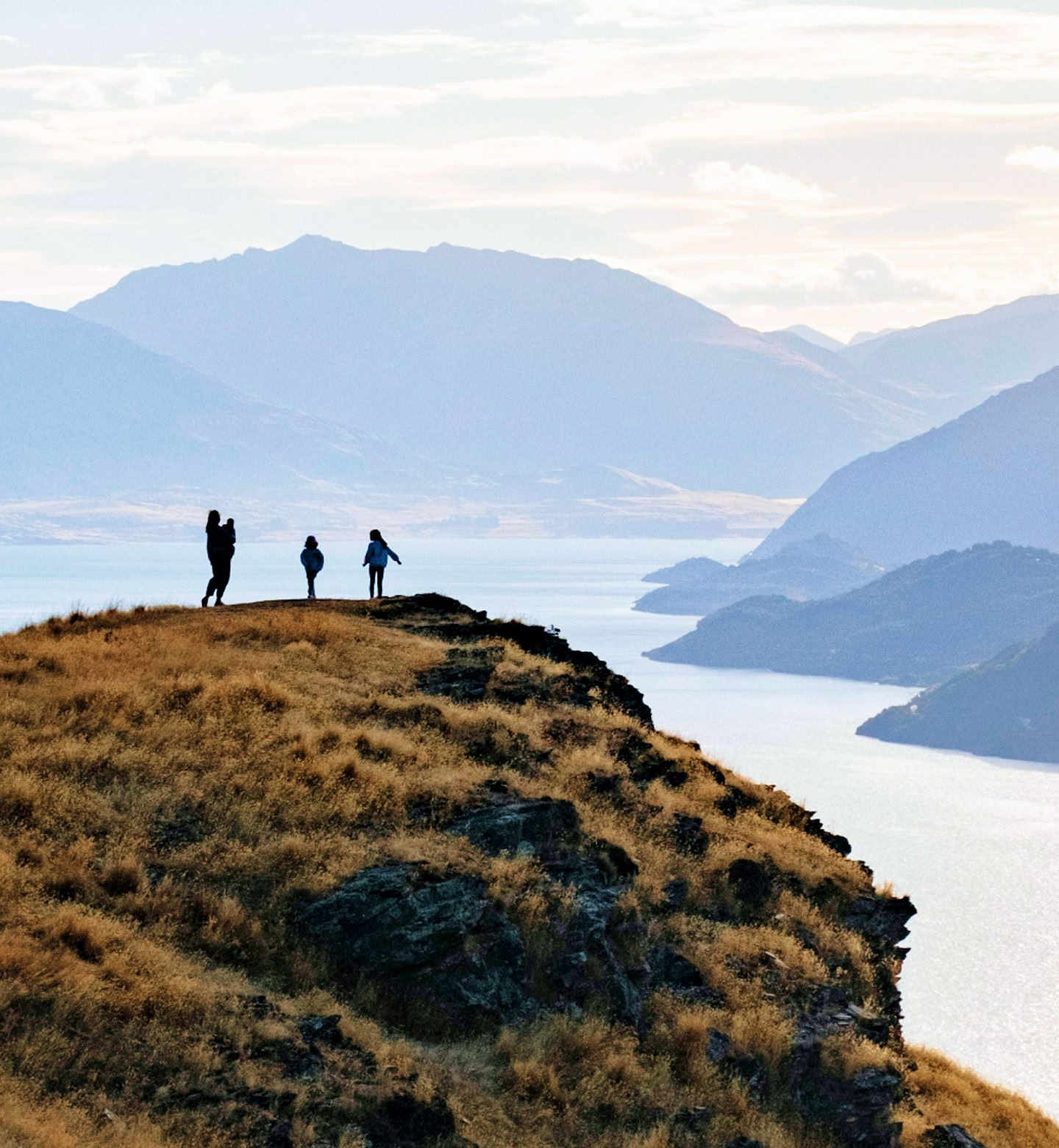 Three people walk along a grassy cliff with lakes and mountains near Queenstown, New Zealand.