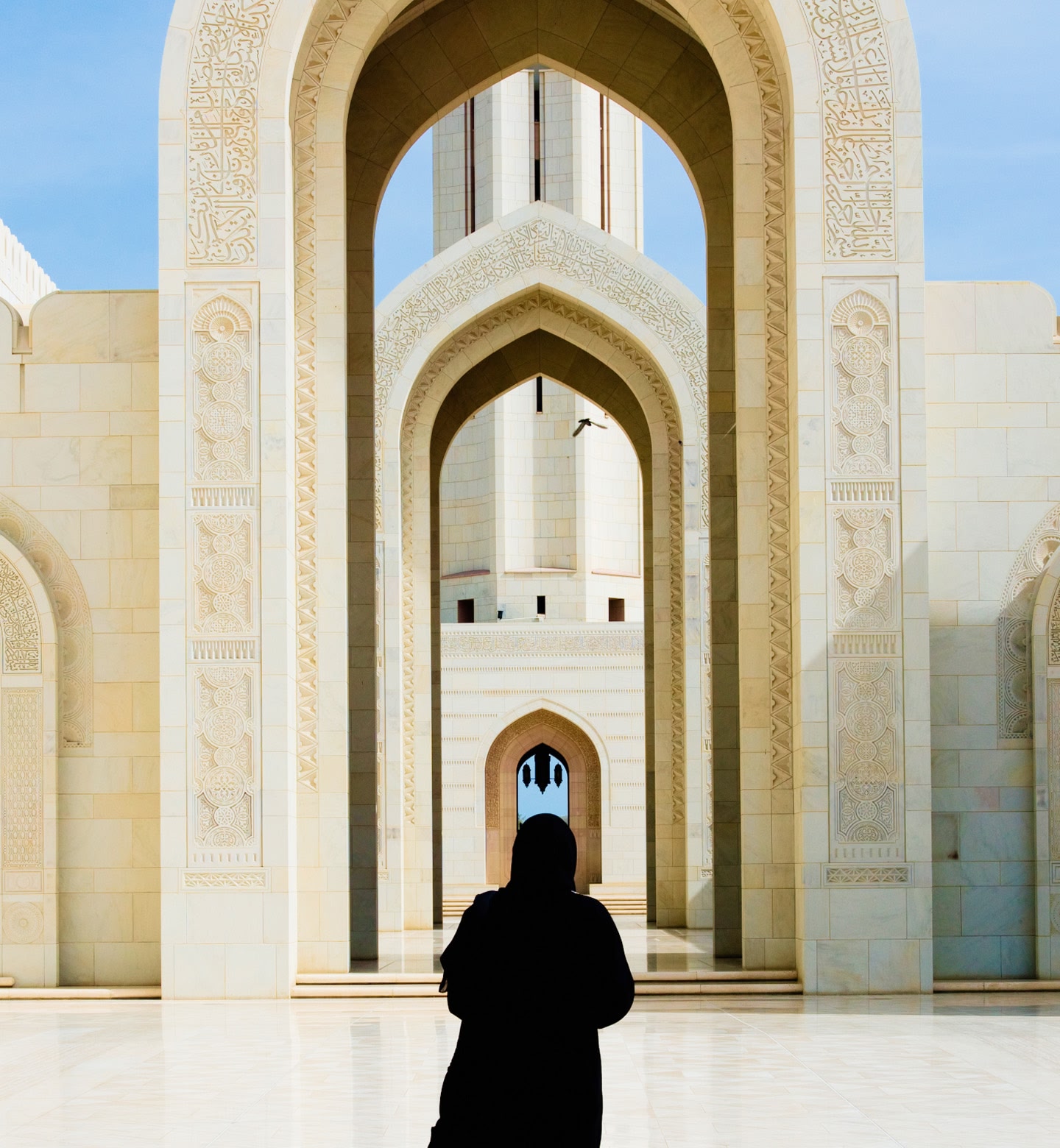 A woman in a headscarf stands in front of a large, white archway at a mosque in Muscat, Oman.