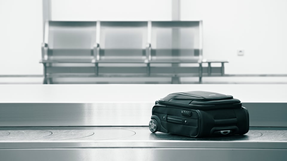 A black rolling suitcase sits on a metal baggage claim carousel in an airport terminal. Empty seating is visible in the background.