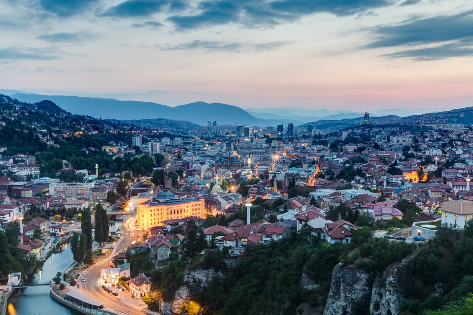 Panoramic view of Sarajevo at dusk, with the Miljacka River, historic red-roofed buildings, and illuminated city landmarks nestled between forested hills and distant mountains.