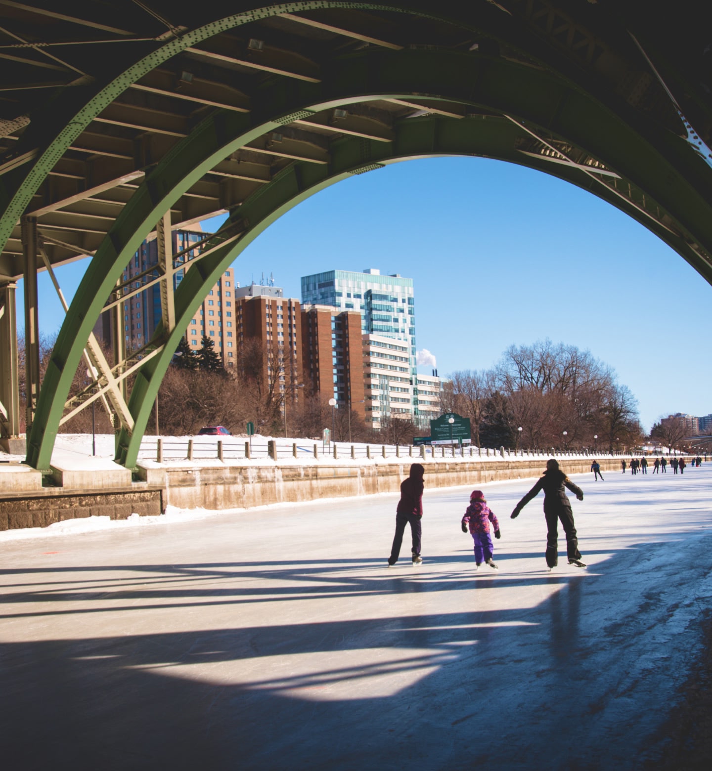 Two adults and a child ice skating underneath a bridge with high rise buildings in the background in Ottawa, Canada