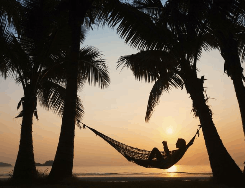 Silhouette of a person in a hammock between two palm trees on a beach, framed by the radiant sunset in the distance, creating a serene tropical paradise scene
