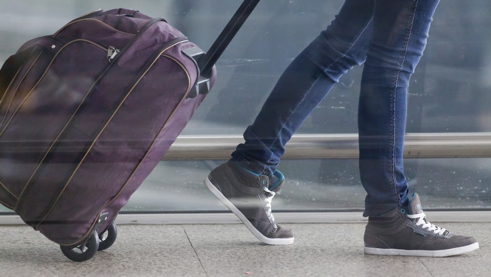 A person in jeans and sneakers walks while pulling a purple suitcase with wheels.