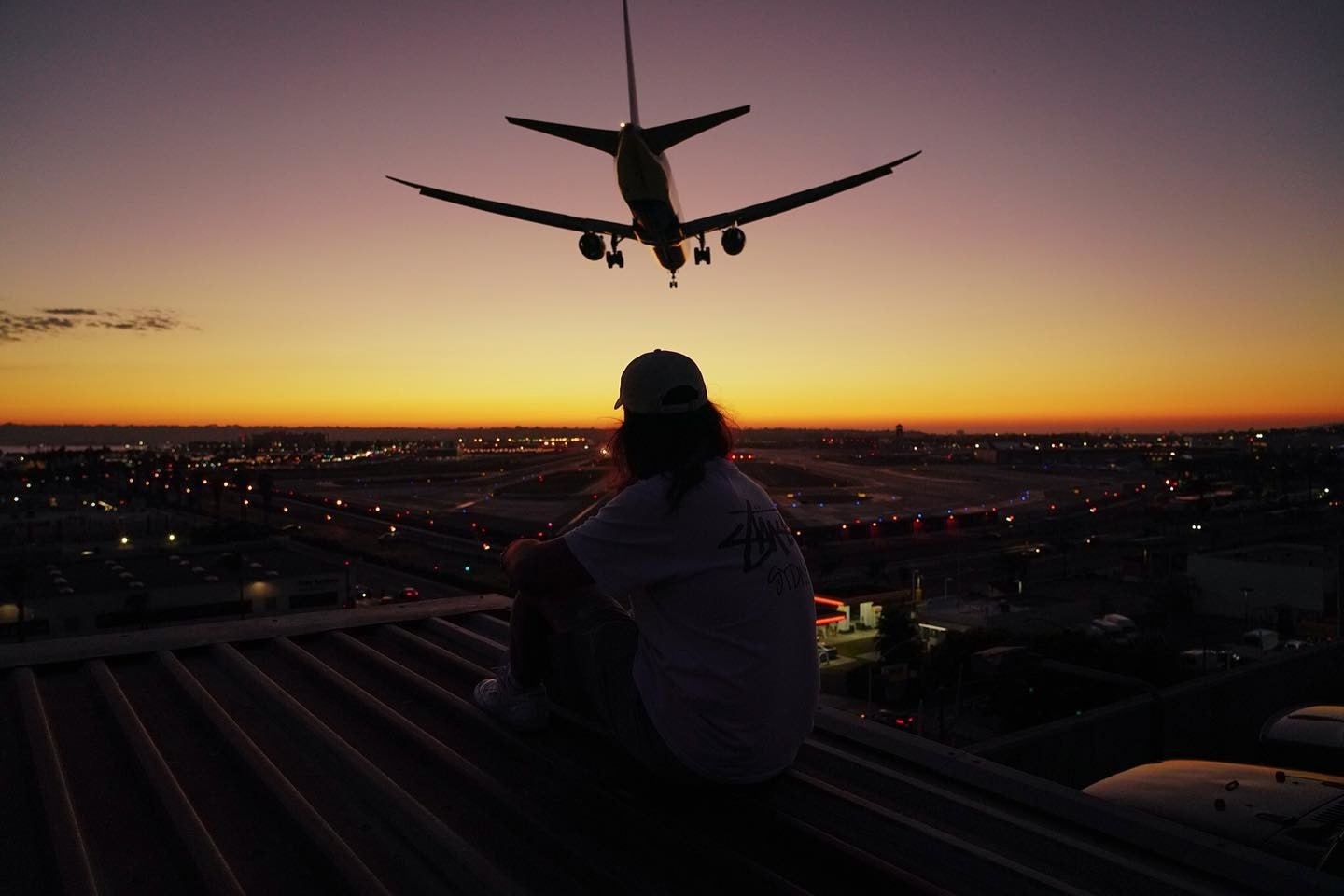 A person sits on a roof by an airport and watches a plane fly over, perhaps contemplating the future of travel.