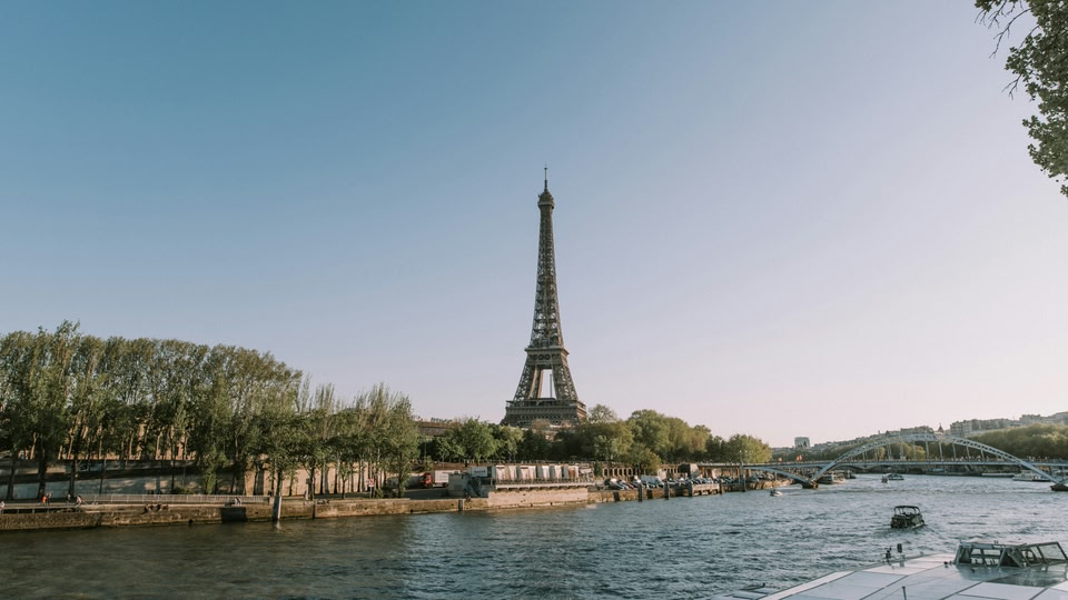 The Eiffel Tower stands tall against a clear blue sky, overlooking the Seine River with its tree-lined banks and a bridge in the distance.
