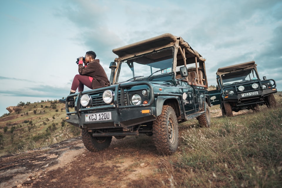 شخص يجلس على مقدّمة مركبة دفع رباعي وينظر إلى الأفق باستخدام منظار