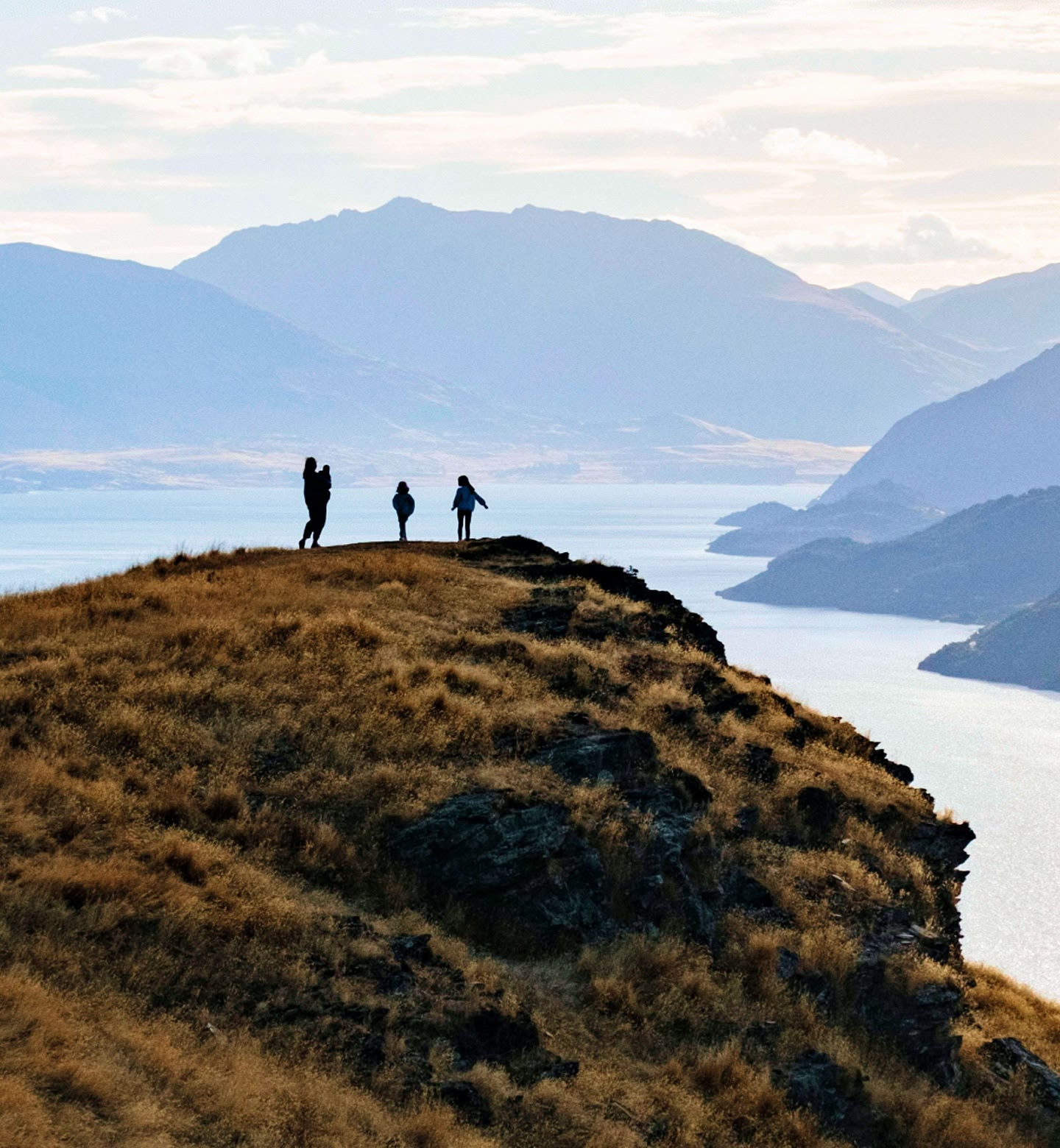 A family stand on a rugged hilltop, silhouetted against a dramatic landscape in Queenstown, New Zealand