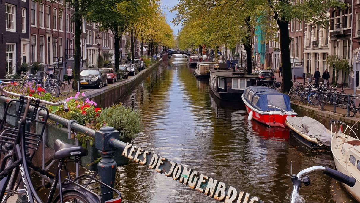Canal in Amsterdam with boats lined up. Cars are parked on the streets either side and people are walking on the pavement.