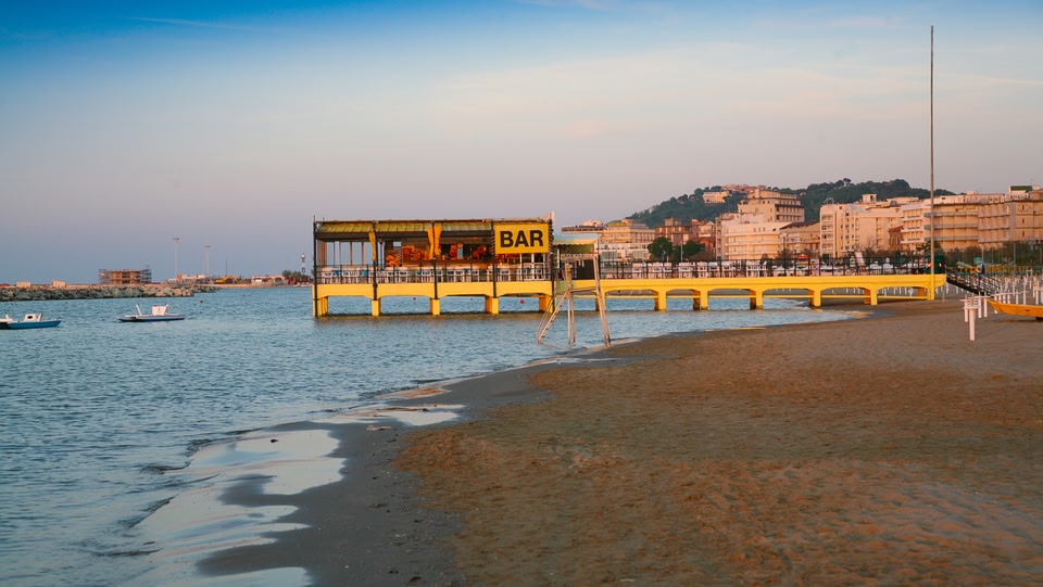 Scena di spiaggia con un bar giallo su un molo che si estende nell'acqua.