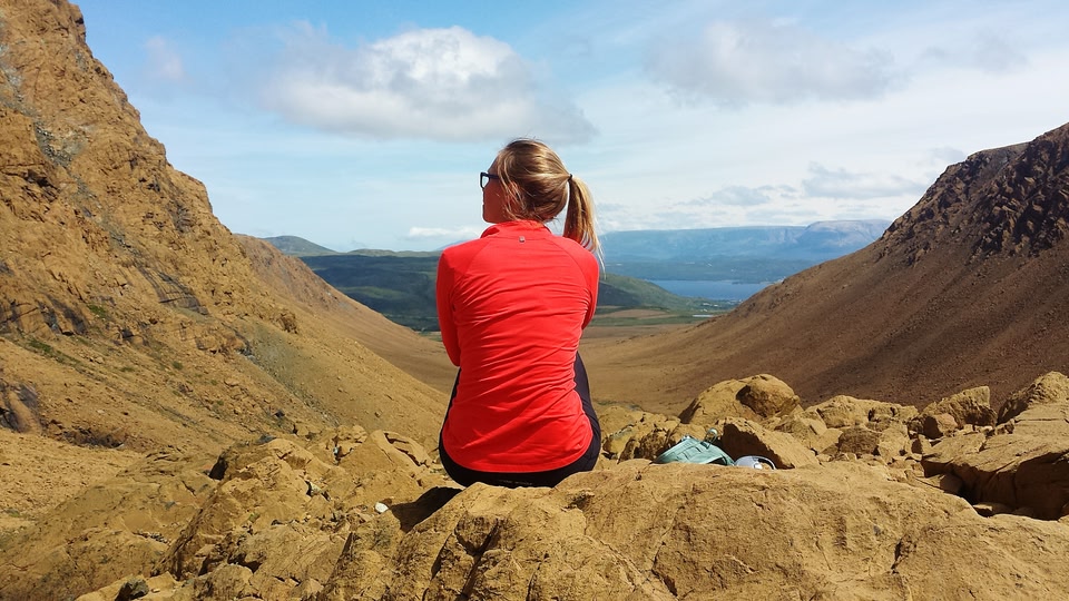 woman in red sweater from behind sitting on a rock at a tourist site in Gros Morne, Newfoundland Canada during the day.