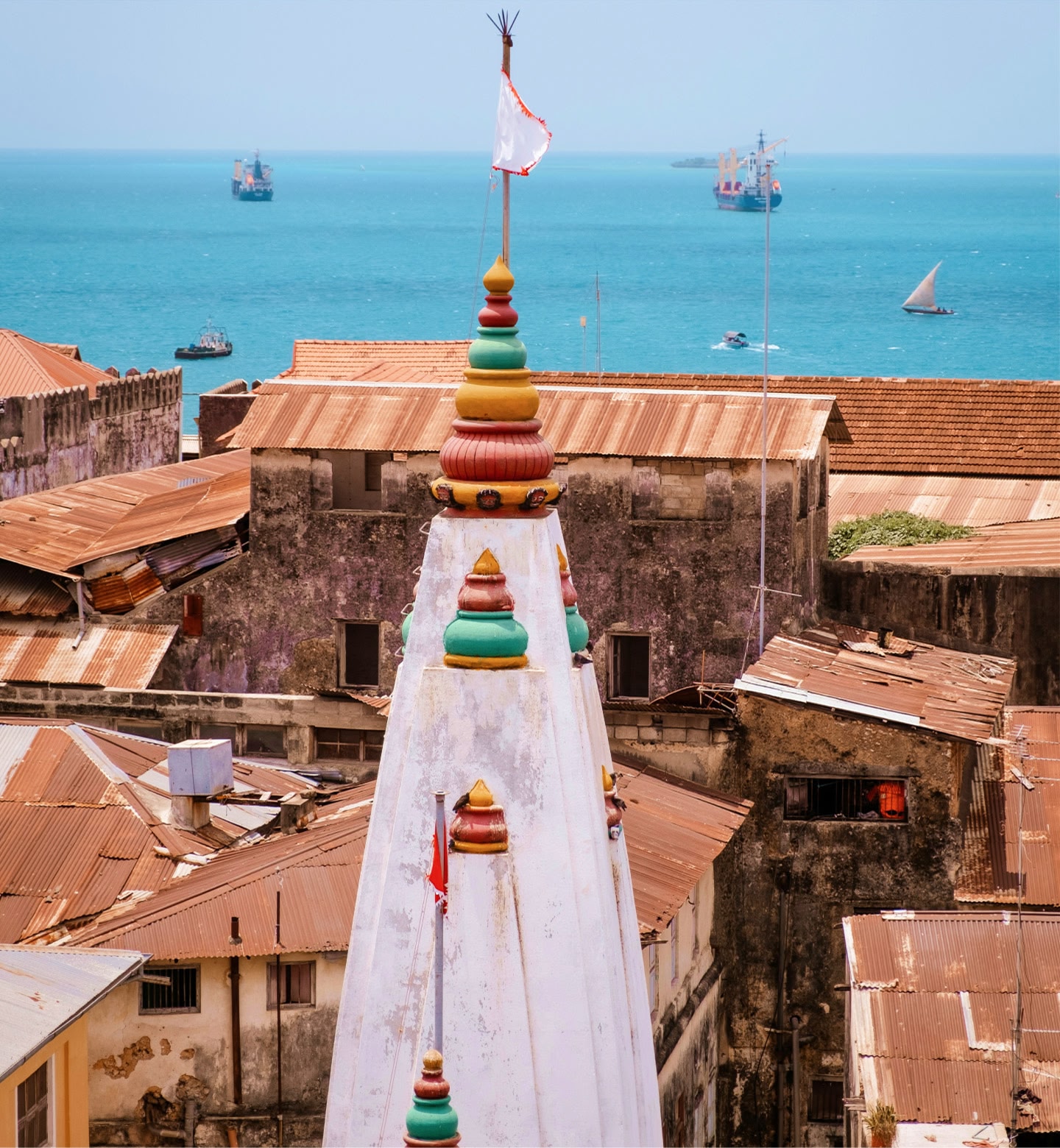The white steeples of a building with colourful tops set against buildings with red roofs and a blue sea.