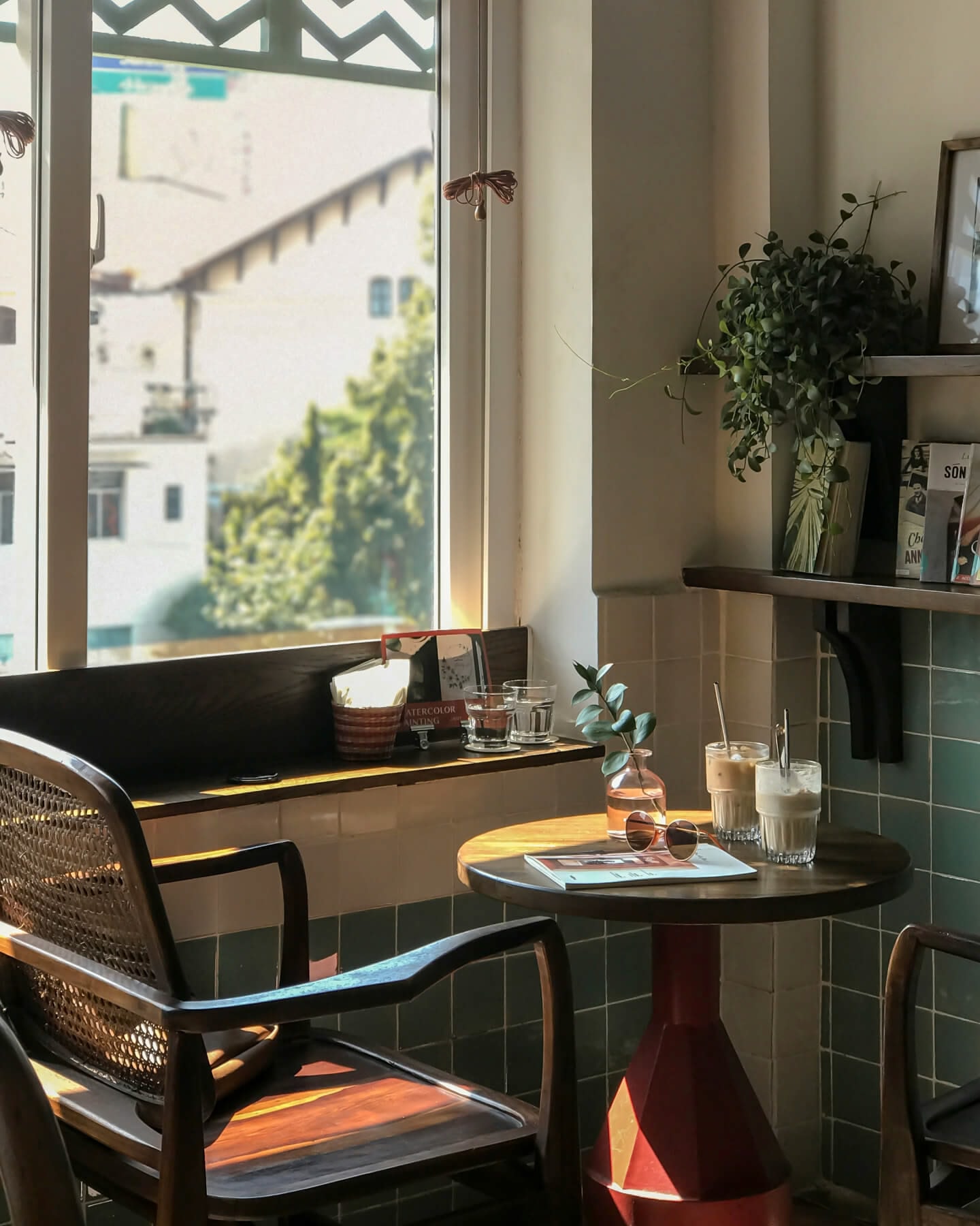 Una pequeña mesa y una silla junto a una ventana, con dos bebidas heladas, una revista y vasos sobre la mesa