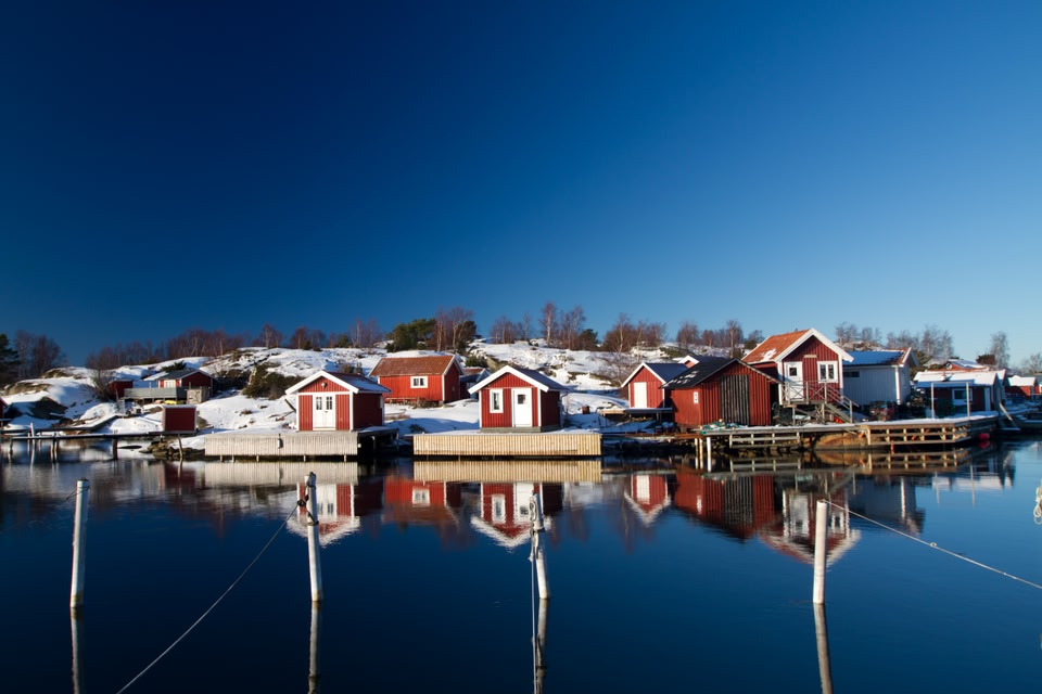 Scenic view of red wooden cabins reflected in calm water on a snow-covered island in the Gothenburg archipelago, Sweden, under a deep blue winter sky.