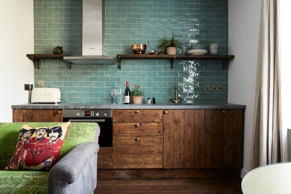 In-room kitchen, with a bottle of red wine and two glasses sitting on the counter. Sofa with a The Beatles cusion in foreground.