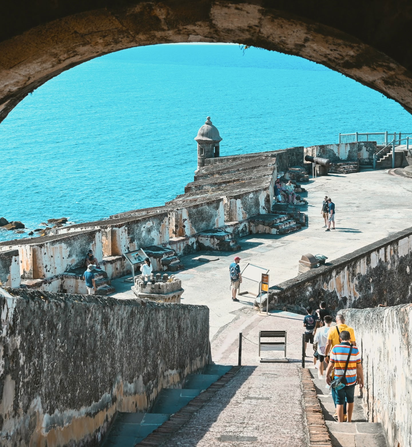 Unos visitantes recorriendo las murallas costeras de un fuerte, con el océano turquesa de fondo en San Juan, Puerto Rico.