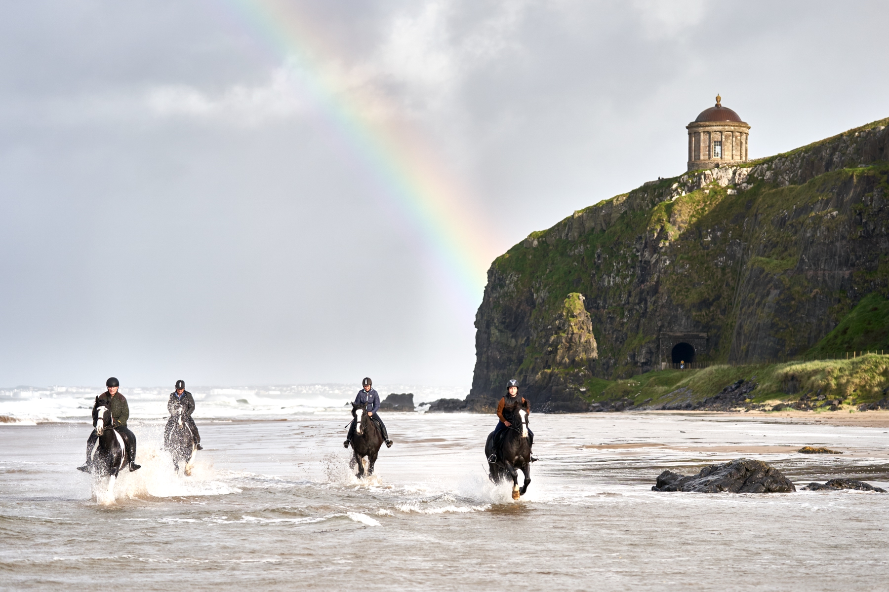 Group of four people riding horses through the waves on Northern Ireland's coast, with a rainbow in the background.