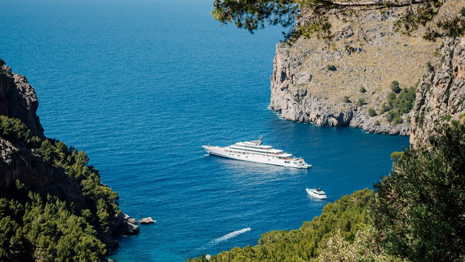 Vista panoramica di una baia con acque blu intenso, scogliere rocciose e uno yacht di lusso.
