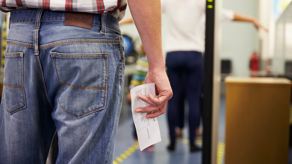 A person wearing jeans and a plaid shirt stands in line at airport security. They are holding a boarding pass in their hand. In the background, another person is going through the metal detector.