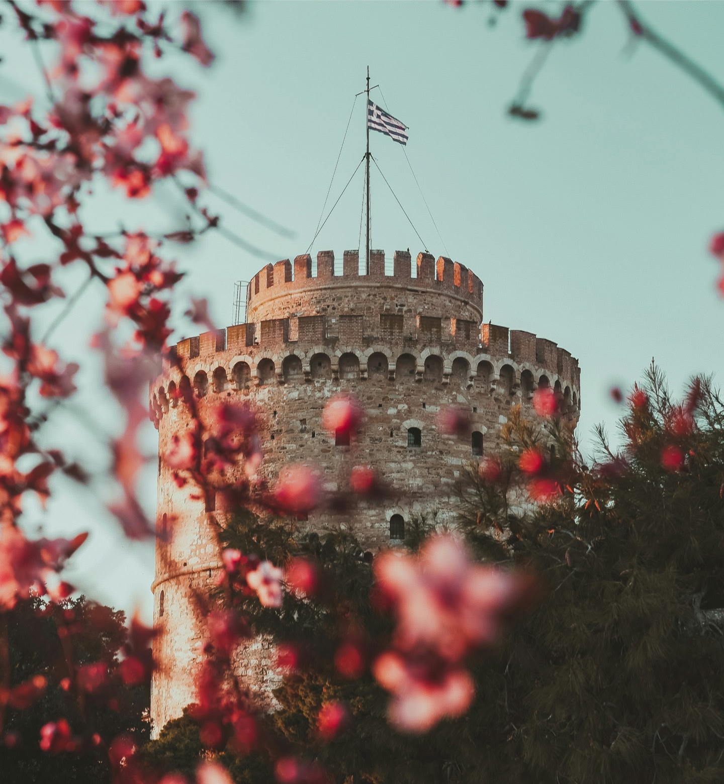 Runder Steinturm mit griechischer Flagge, eingerahmt von rosa Blüten, in Thessaloniki, Griechenland