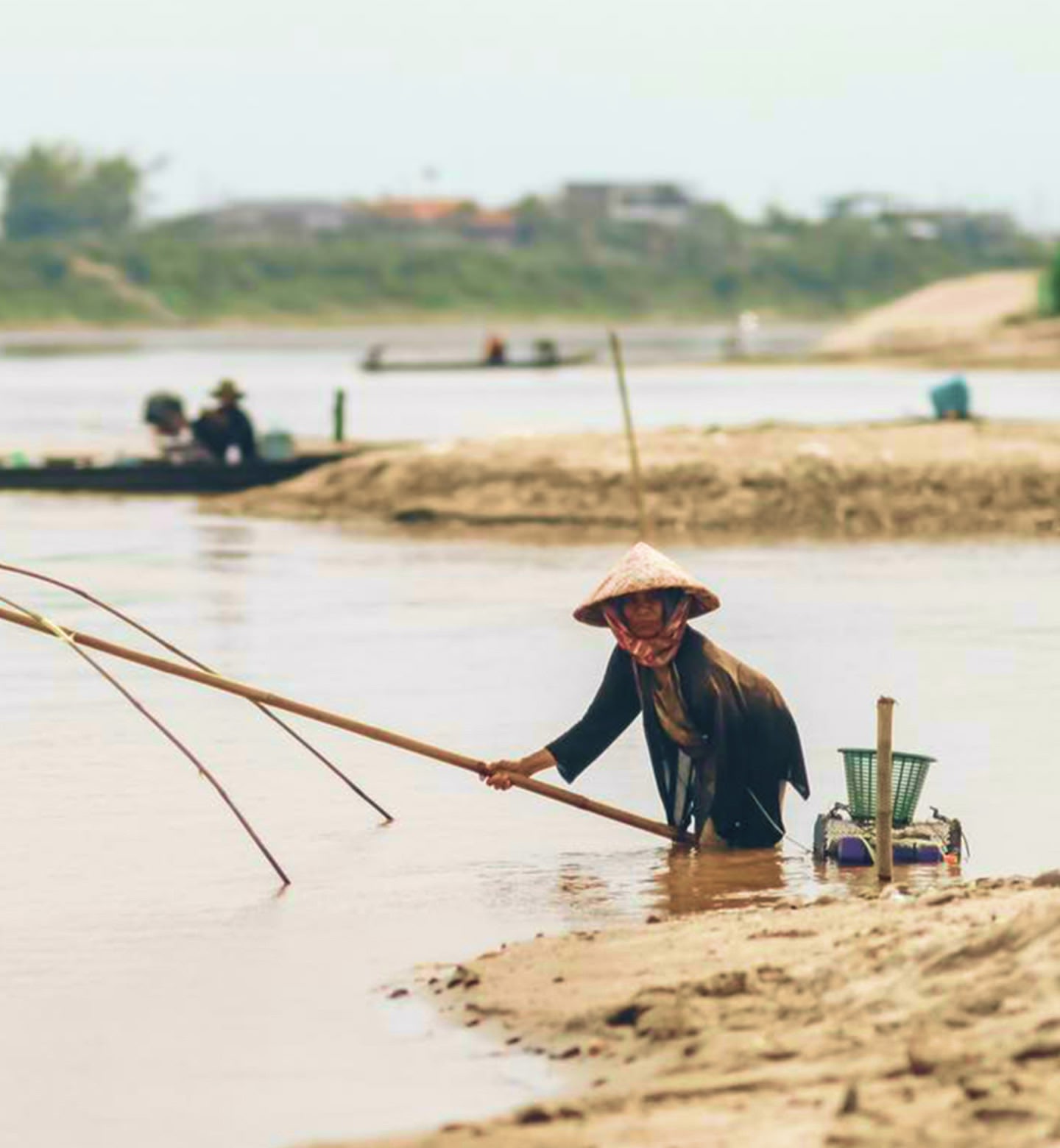 ラオスのヴィエンチャンで、円錐帽をかぶった漁師が長い竿で体を支えて川の中に立っている。