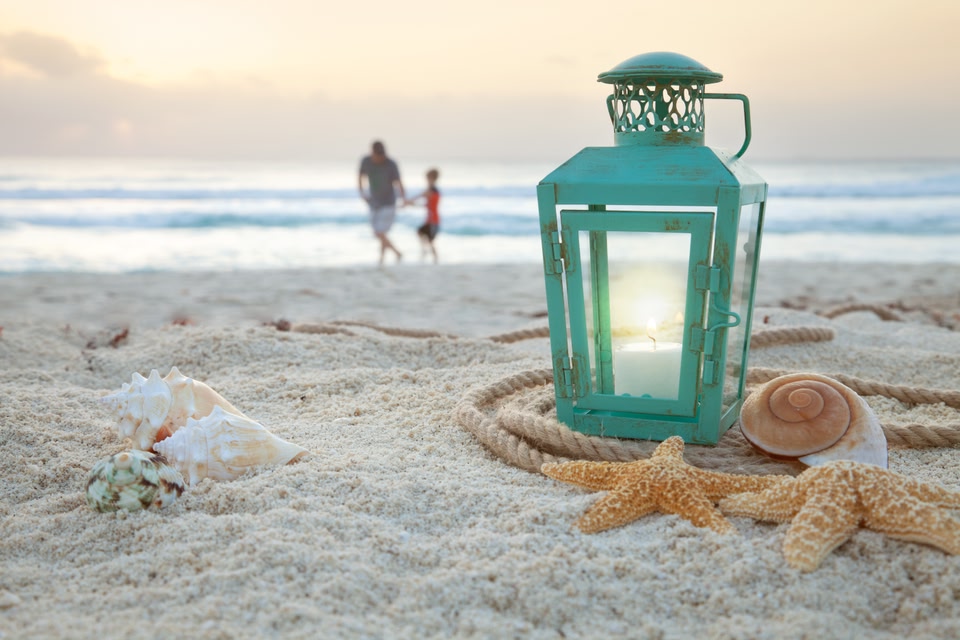 Lantern and seashells on a sunset beach, with a father and sun splashing in the waves in the background.