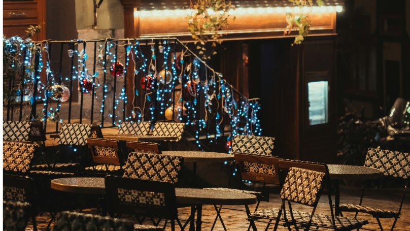 A picture of patterned chairs and round tables of a café laid out on a street at night with a row of blue fair lights wrapped around a rail