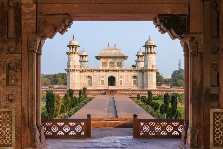 The Baby Taj in Agra viewed through an intricately carved red sandstone gateway, with a formal garden pathway lined with trees and shrubs leading to the white marble tomb flanked by four minarets.