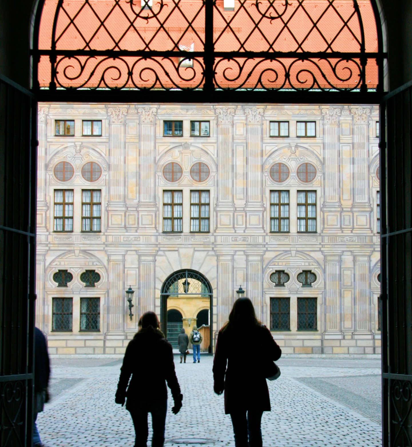 Two women walk in the courtyard of Munich Residence, Germany, viewed from an intricate metal gateway