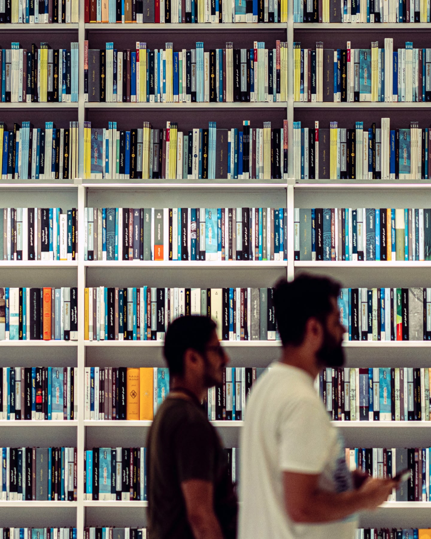 Two men walk past a ceiling-height set of white bookshelves, full of books.