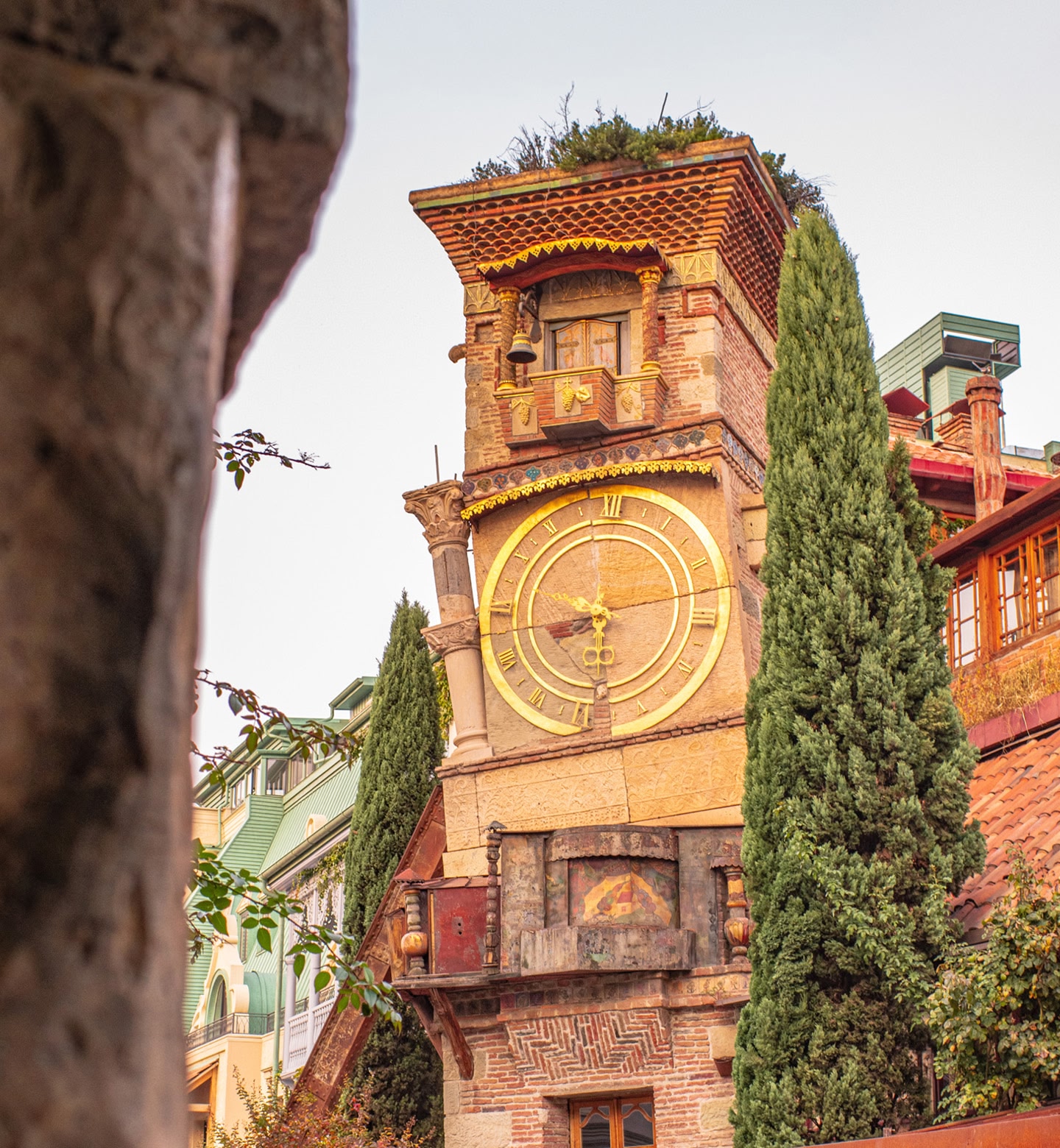 Close up shot of a quirky, leaning clock tower flanked by green trees in Tbilisi, Georgia.