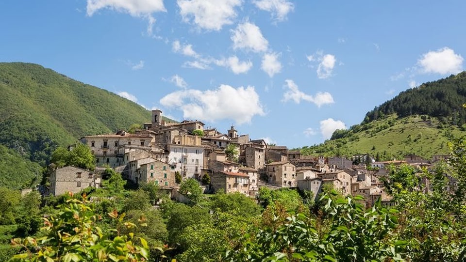 A scenic view of a traditional Italian village with stone buildings, situated on a hillside surrounded by lush green mountains and a blue sky with scattered clouds.