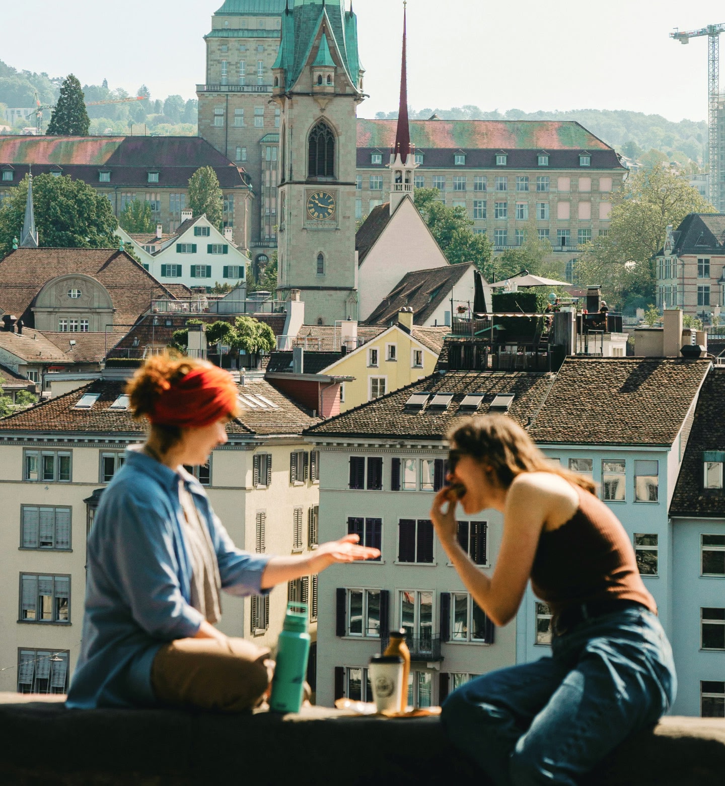 Two woman talk and eat while sitting on a balcony overlooking buildings in Zurich, Switzerland.
