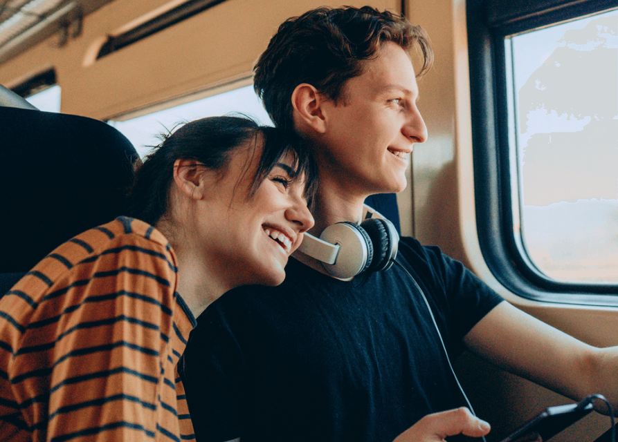 Two people sitting on a train looking out the window. One person is leaning their head against the shoulder of the other person.