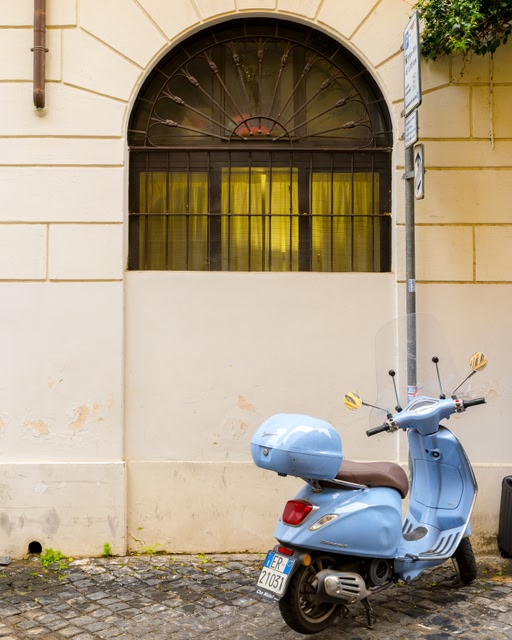 A light blue vespa parked on the street next to a curved window.