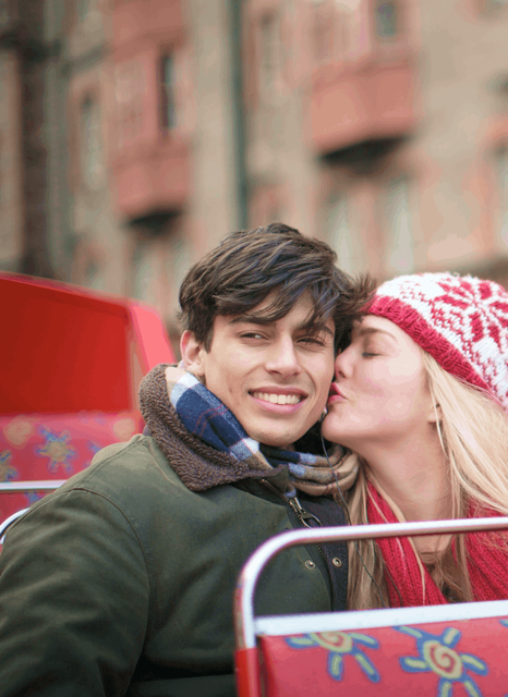 A close up shot of a young blonde woman wearing a hat, kissing a dark haired smiling male on the cheek, while sat on the top of a London open top bus.