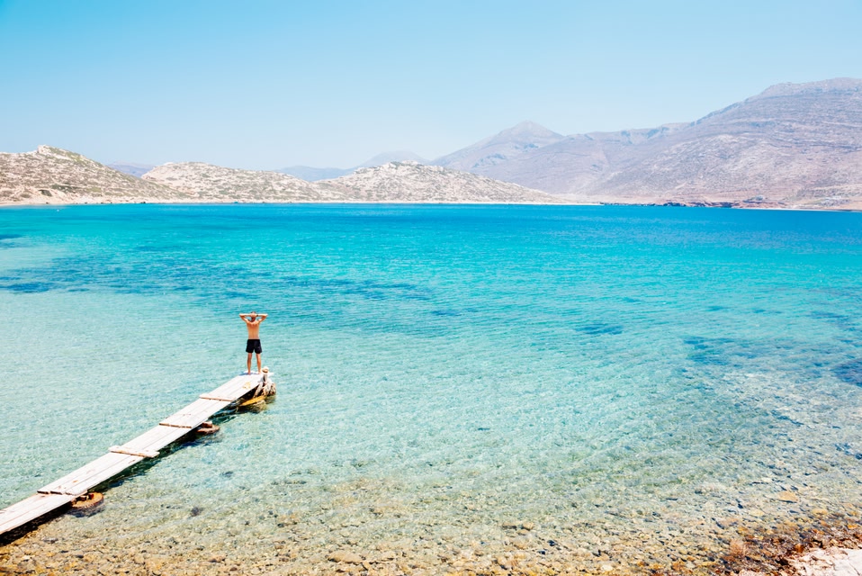 Man on a boardwalk gazes at the turquoise sea and surrounding cliffs in Amorgos, Greece